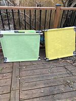 Two chaise lounges placed side by side outdoors on wooden decking, one green and one yellow, shown folded or standing upright.