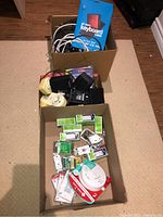 Two cardboard boxes on carpeted floor; one box contains black and white electrical cords, some small electronics, and cases; second box contains electrical outlets, faceplates, and a package containing a First Alert smoke and CO detector.