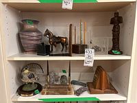 Two shelves holding various decorative items including a large pottery vase, copper horse figurine, wooden bookends, Inuit carving, glass ink stand, hand-painted bird figurine, and wooden totem statue.
