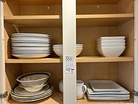 Two shelves of assorted Crate and Barrel dishes, including white round plates, bowls, square plates, and mugs.
