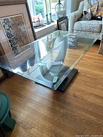 Glass top coffee table shown from an angled top view, displaying the beveled edges and transparency of the glass surface. Faux wood base can be seen underneath.