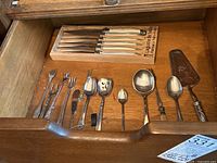 Opened drawer revealing set of six steak knives and various metal serving utensils including forks, spoons, and a cake server.