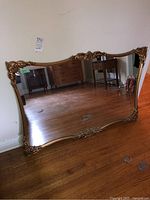 Full view of large rectangular ornate mirror resting against wall, clear reflective surface with some floor marks.