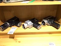 Three vintage Hanimex movie cameras displayed on a wooden shelf, showing different body colors and lens assemblies.
