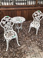Photo showing full set of white metal patio furniture: two chairs and a round table placed outdoors on a bed of dried leaves and stone pavers. Visible paint flaking and wear on all pieces.