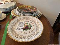 Footed plate with gold trim and floral fruit center front, stacked plates including Norman Rockwell collector plate in background