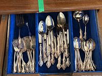 Full drawer of silverware including forks, knives, spoons, serving spoons, and ladles, arranged in separate compartments lined with blue velvet.