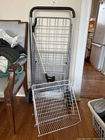 Foldable metal shopping cart frame with two black wheels, black handle, and separate white wire basket on wood floor next to a chair.