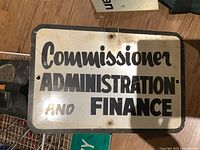 White and black metal sign reading 'Commissioner Administration and Finance' showing rust and age marks