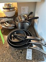 Photo showing assortment of non-stick Rock frying pans stacked on counter with stainless steel saucepans with black handles and glass lids.