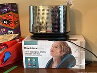 Front view of Bodum perforated stainless steel toaster on top of Brookstone massager box, showing the toaster's reflective surface and perforated design.