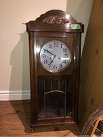 Front view of the vintage wooden wall clock showing carved wooden detail on top, white clock face with black numbers and hands, and visible pendulum through bottom glass panel.