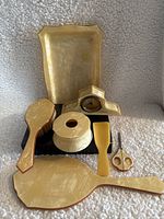 Image showing full vintage vanity set items arranged on a white textured fabric background, includes mirror, brush, tray, covered box, clock, scoop, and scissors.