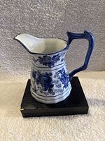 Front view of Chinese blue and white floral porcelain pitcher on black stand, angled spout, and handle, showing detailed floral motifs.
