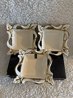 Three white square porcelain dishes with ornate gold trim edges and handles stacked on a black box on a textured cream fabric.