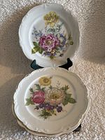 Three white porcelain plates with colorful hand painted flowers, displayed on a fabric background, showing detailed flower patterns and scalloped rims.