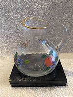 Full view of the clear glass pitcher showing floral hand-painted decorations with gold trim on the rim, curved handle, and ribbed glass body.