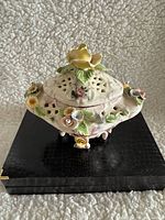 Image showing side view of a white porcelain decorative box with hand-painted pink accents, pierced lid, three-dimensional yellow, pink, and white flowers, green leaves, and three small legs.