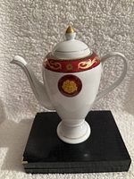 Front view of white coffee pot with red and gold Medusa design, showing spout and handle