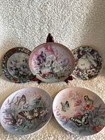 Five decorative collector plates displayed on beige fabric background, one mounted on wooden display stand in center.