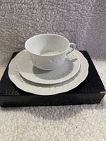 Porcelain cup, saucer, and cake plate set displayed stacked on a black box with visible embossed floral pattern on each item.