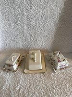 Three porcelain butter dishes in a row on a textured fabric background, showing top and front views. The middle dish is ivory with gold trim and visible crazing. The side dishes are floral patterned.
