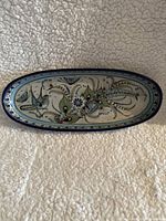 Top view of oval ceramic platter showing hand-painted birds, butterflies, and floral designs in blue, green and brown with blue border rim.