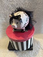 Front and top view of pink ceramic piggy bank dressed in black tulle tutu and polka dot bow on black and white striped round box with pink lid