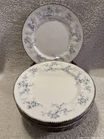 Stack of 10 white porcelain dinner plates with blue floral pattern and silver trim visible around edges.