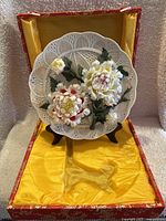 Decorative porcelain plate with dimensional pink and white flowers on yellow fabric inside original red box with stand.