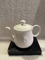 Front and side views of white porcelain teapot with raised cameo medallion on side, placed on a black textured stand, against a textured light background.