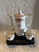 Porcelain coffee pot, creamer, and sugar bowl on black base, white with pink floral pattern and gold trim.