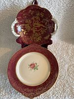 Two red and gold porcelain serving plates with detailed floral and bird designs. One plate has handles and a deep red surface with gold birds and florals. The other plate is larger, with a red and gold rim and central pink rose on white porcelain.