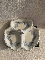 Top-down view of the divided porcelain dish showing blue floral decoration on each section, gold trim on scalloped edges, and a central handle.