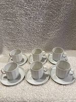 Six white porcelain coffee cups placed on their matching saucers, arranged in two rows on a textured grey background.