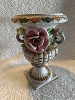 Front view of the ceramic vase showing a large pink rose sculpture, detailed leaves, ornate side handles, and ribbed body with orange-green brush strokes