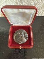 Front view of bronze medallion with classical figurative winged female design resting in red velvet case with white satin lid lining.