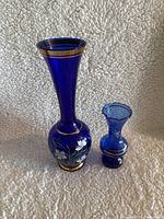 Two cobalt blue decorative glass vases with gold trim and white hand-painted floral accents, different heights with flared neck and ruffled rim.