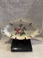 Full view of cream-colored porcelain serving platter with pink rose decorations and scalloped edges on a red stand.
