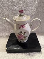 Front angled view showing the spout, handle, and floral design on white porcelain teapot with rose finial lid.
