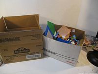 First image showing a large cardboard box filled with various LEGO-style base plates in green, blue, and brown along with assorted blocks and pieces.