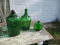 Three green glass bottles in varying shapes displayed on a worn wooden table outdoors.