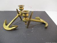 Two brass anchor-shaped candle holders displayed side by side, showing front and side views.