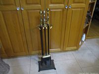 Full view of four fireplace tools in stand, placed on tile floor in front of wood cabinets.