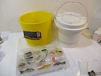 Wide shot of a yellow floating minnow bucket, white bucket with handle, and open plastic tackle box with assorted fishing lures inside.