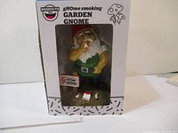 Front view of the BigMouth Inc. gNOme Smoking Garden Gnome figurine inside box, showing the gnome smoking a cigarette with accessories visible.