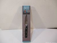 Front view of the kitchen knife in packaging showing blade and handle aligned inside the box that is mostly grey and teal with Jamie Oliver branding.