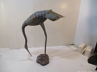 Side view of metal coppertone bird sculpture on a plain background showing detailed feathers and long curved neck.