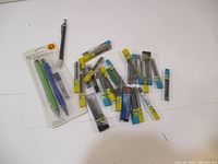 Lot of 3 mechanical pencils and multiple tubes of lead refills scattered on white surface.