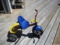 Front and side view of the kids tricycle showing blue seat, yellow platform, red frame, and tire details on wooden deck.